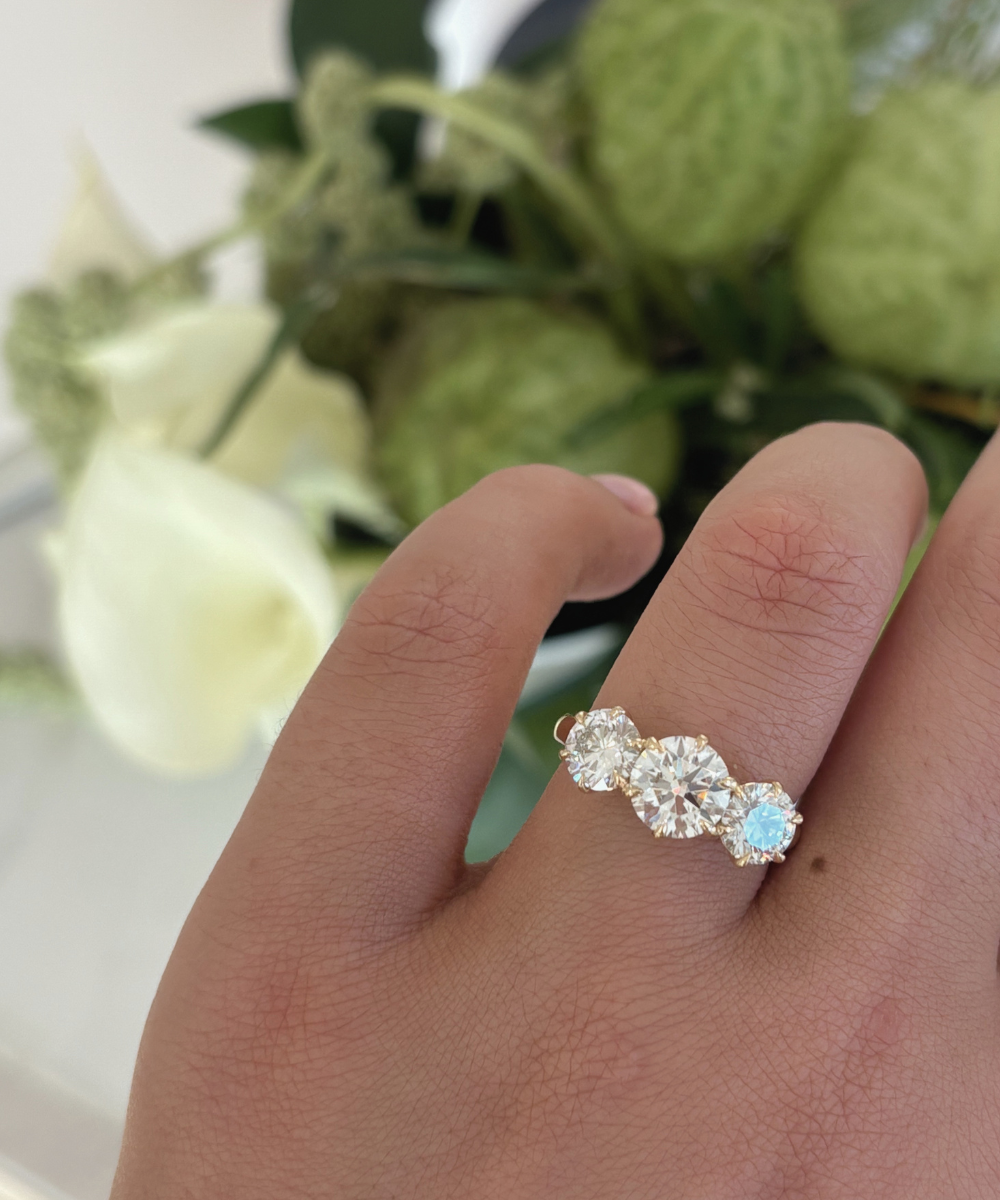Portia trilogy engagement ring worn on finger with white calla lilies highlighting sparkle of round lab grown diamonds