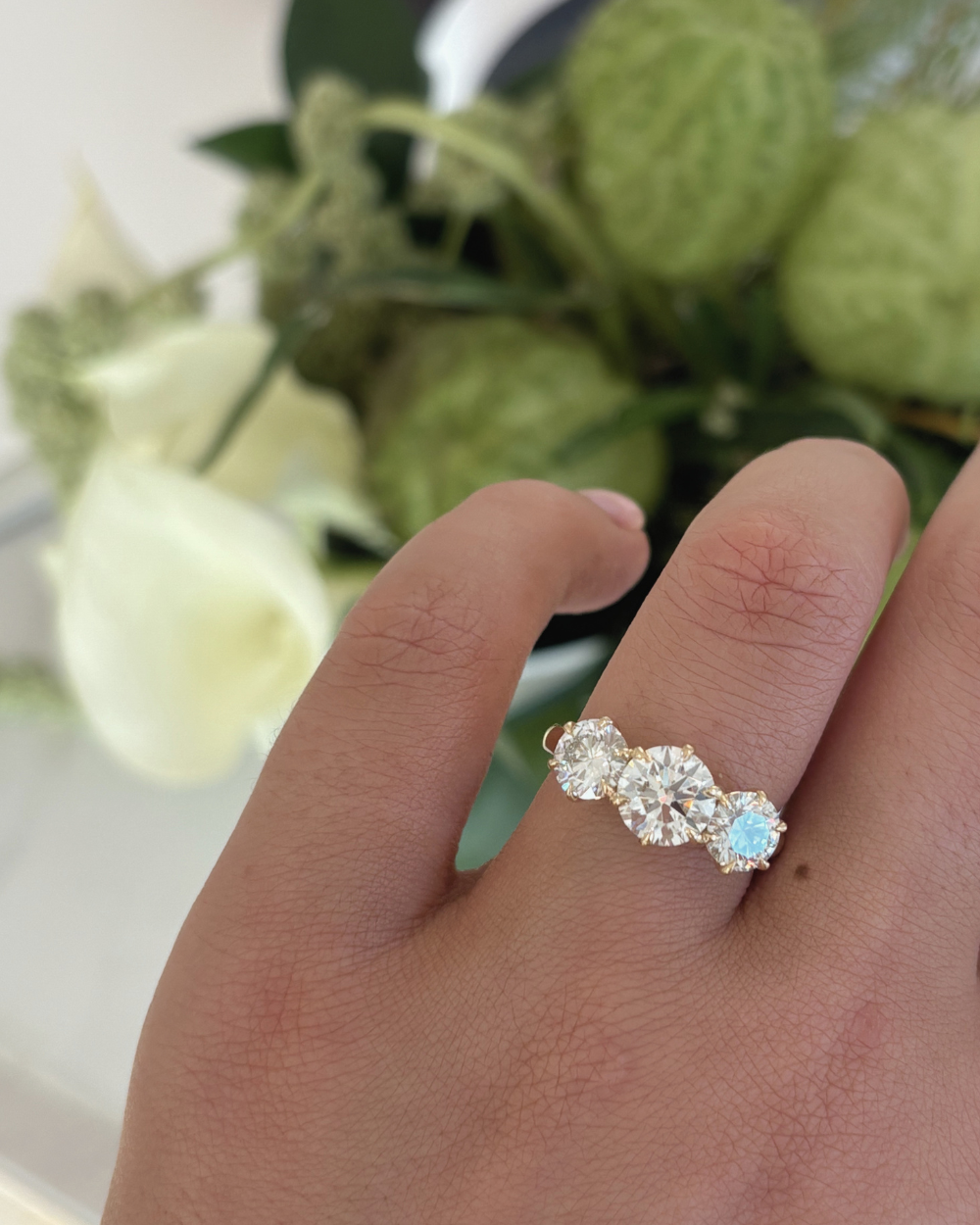 Portia trilogy engagement ring worn on finger with white calla lilies highlighting sparkle of round lab grown diamonds
