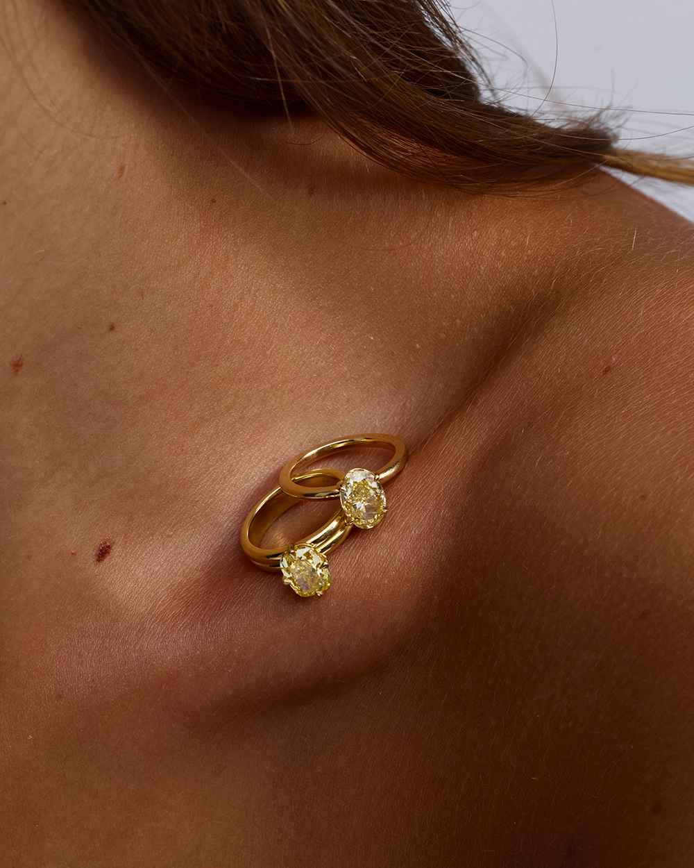Two oval yellow diamond solitaire rings resting on bare shoulder showing scale