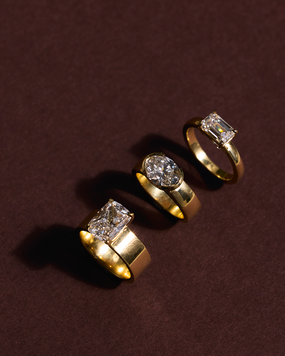 Three 18ct yellow gold engagement rings with cushion, oval, and emerald cut diamonds on burgundy background
