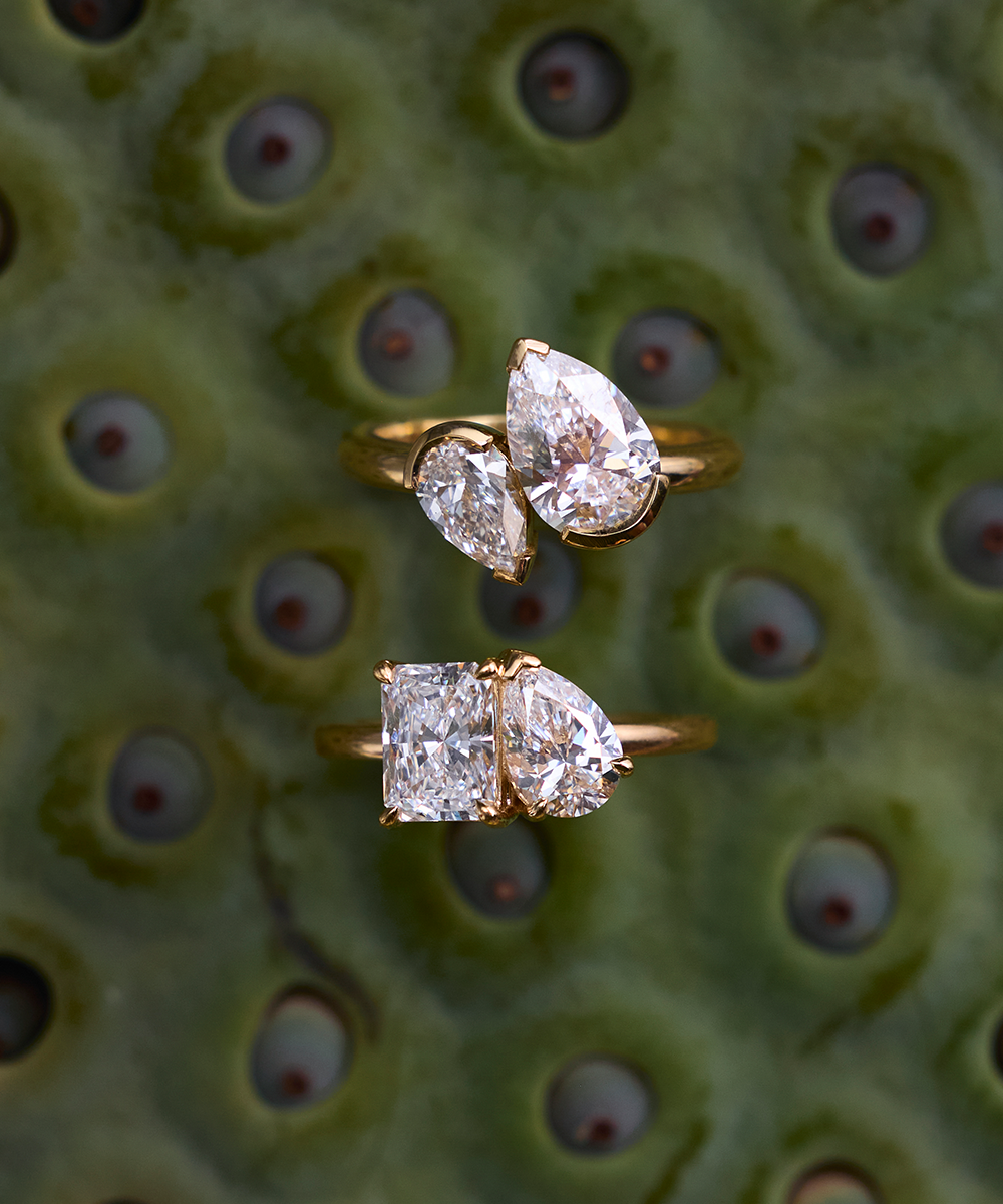 Two Nova rings on green textured surface featuring radiant-pear and pear-marquaise diamond pairs in yellow gold