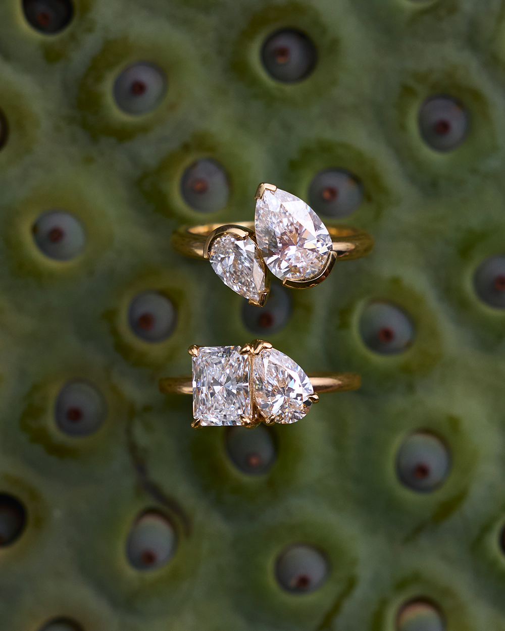 Two Nova rings on green textured surface featuring radiant-pear and pear-marquaise diamond pairs in yellow gold