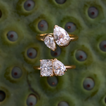 Two Nova rings on green textured surface featuring radiant-pear and pear-marquaise diamond pairs in yellow gold