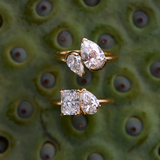 Two Nova rings on green textured surface featuring radiant-pear and pear-marquaise diamond pairs in yellow gold
